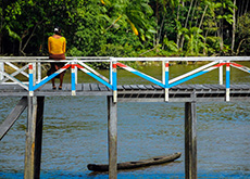 Fotografia mostra uma ponte de madeira, com as laterais pintadas nas cores branca, azul e vermelha. Em baixo da ponte, passa um rio. Sentada na proteção lateral, de costas para nós, uma mulher observa a vegetação no fundo da imagem. 